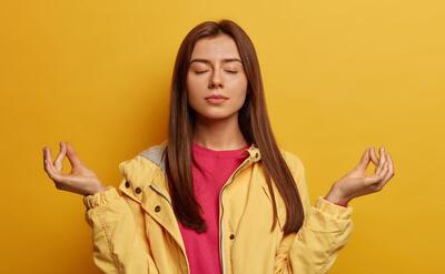 Relaxed dark haired young woman with healthy skin, makes zen mudra gesture, tries to calm down after hard day, breathes deeply and practices yoga, wears windbreaker, isolated on yellow wall