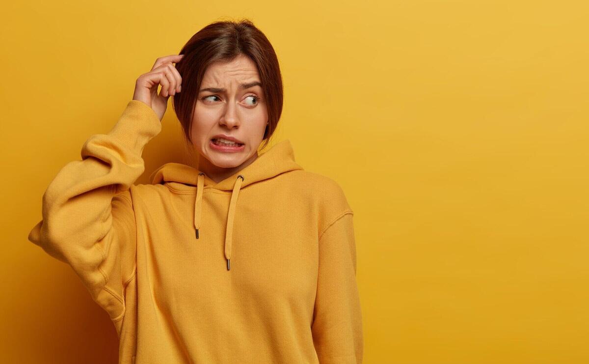 Unsure doubtful woman scratches head, has amnesia, makes tough decision, looks troubled aside, stands puzzled, clenches teeth, dressed in hoodie, poses against yellow wall, empty space for text