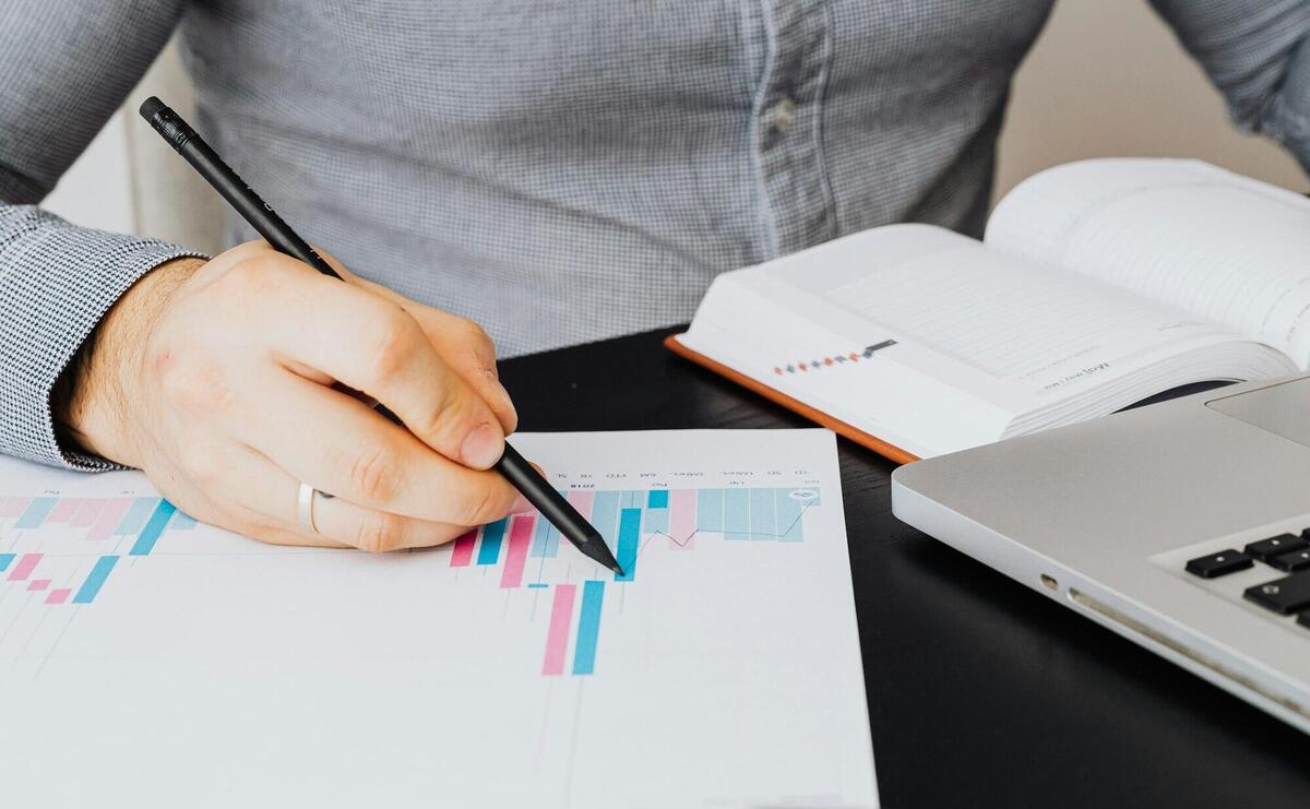 A business analyst examines a graph on paper with a laptop and notebook indoors.