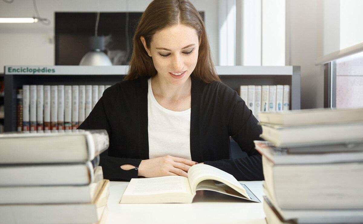 Portrait of beautiful young brunette female professor wearing black jacket reading manual or textbook, smiling, preparing for lecture in university, sitting at library in front of stacks of books