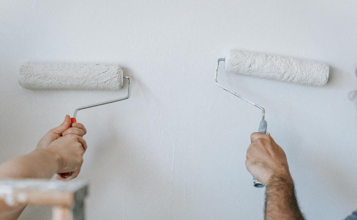 Close-up of two individuals painting a wall with paint rollers indoors.