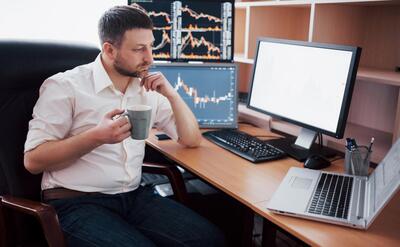 Young businessman is sitting in office at table, working on computer with many monitors,diagrams on monitor. Stock broker analyzes binary options charts.Hipster man drinking coffee,studying