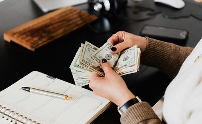 Unrecognizable elegant female in sweater counting dollar bills while sitting at wooden table with planner and pen