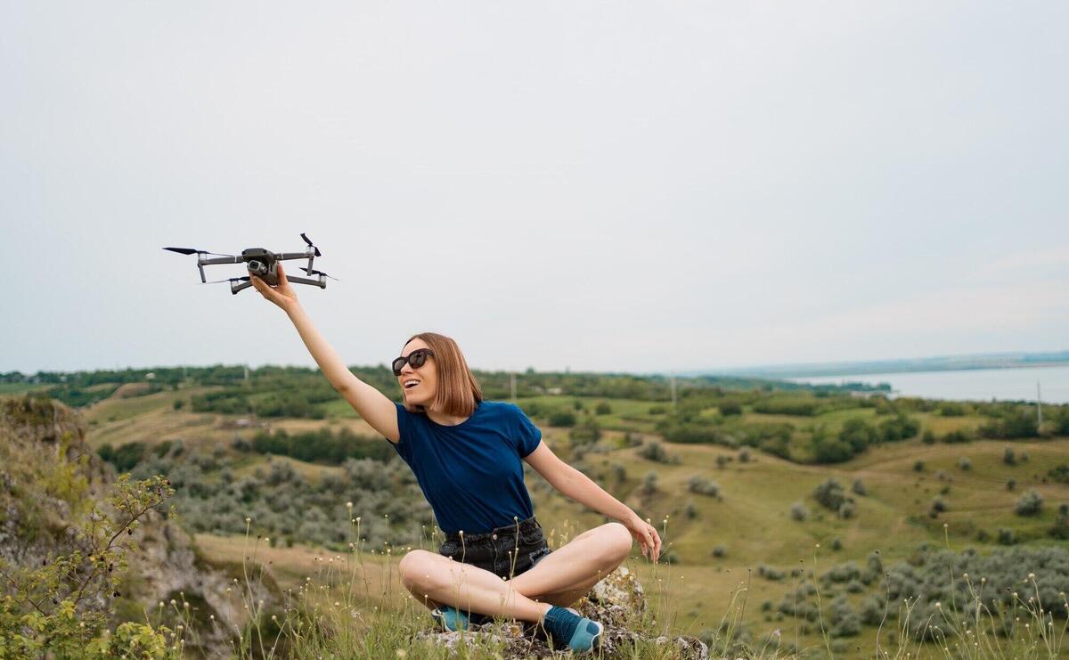 A Caucasian woman with a drone in her hand, sitting on a green rocky hill with sky in background