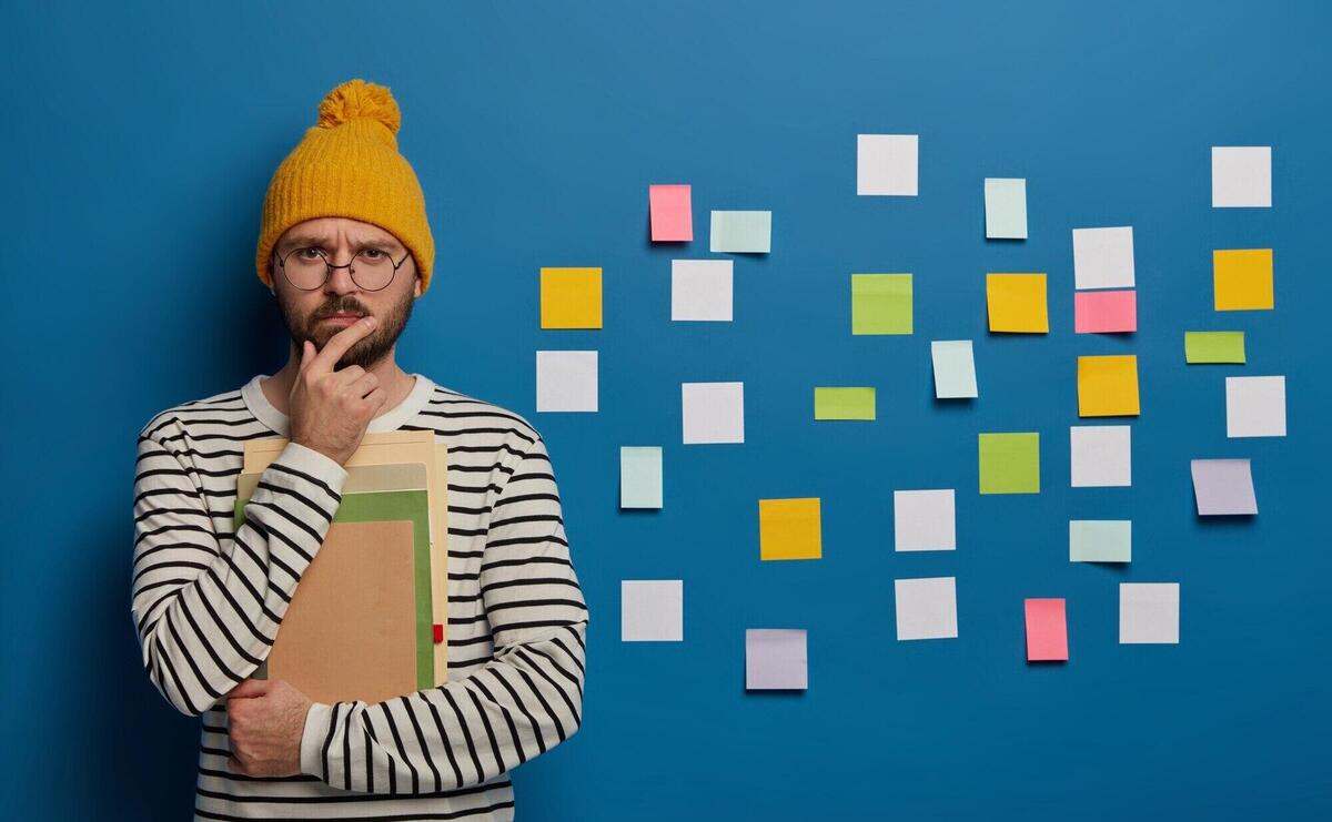 Serious mentor leader thinks on creative ideas, holds chin and looks straightly at camera, wears transparent round glasses, stands with notepads