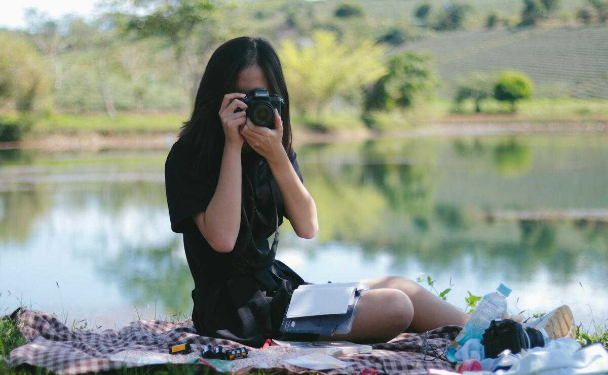 woman using camera while sitting on sheet on grass beside the body of water