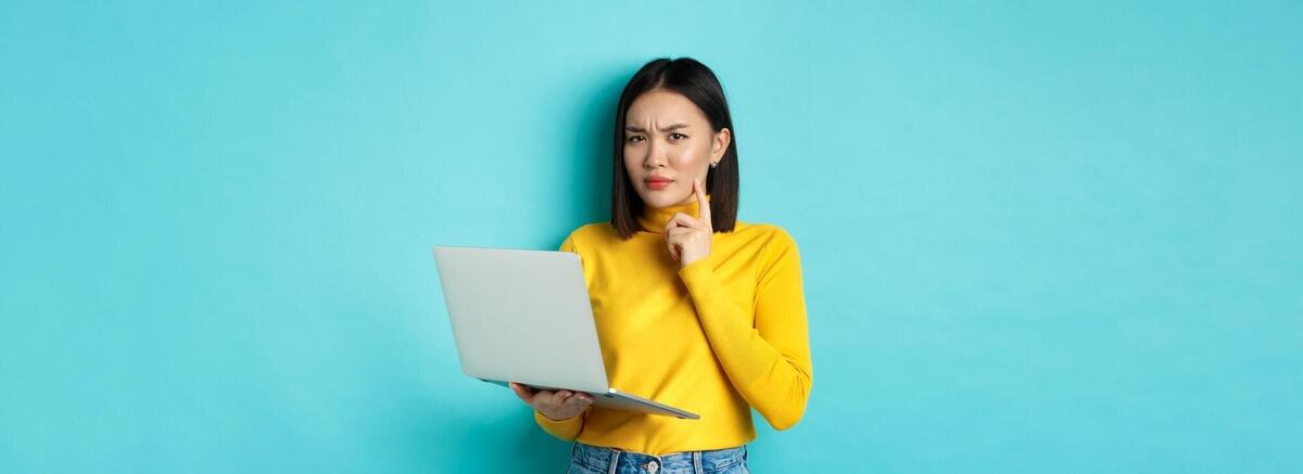 Serious looking asian woman working on laptop and thinking frowning at camera solving problem at wor