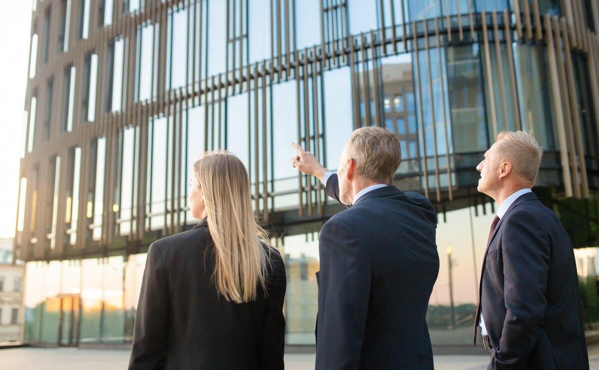 Group of business partners in formal suits pointing at office building, meeting outdoors, discussing real property. Back view. Commercial real estate concept