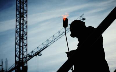 Silhouette of a construction worker using a blowtorch at a building site against a crane-filled skyline.