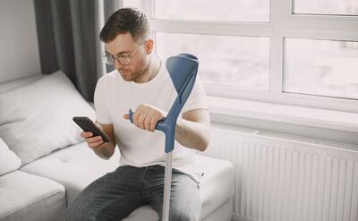 Young man with broken leg at home. Using smartphone.