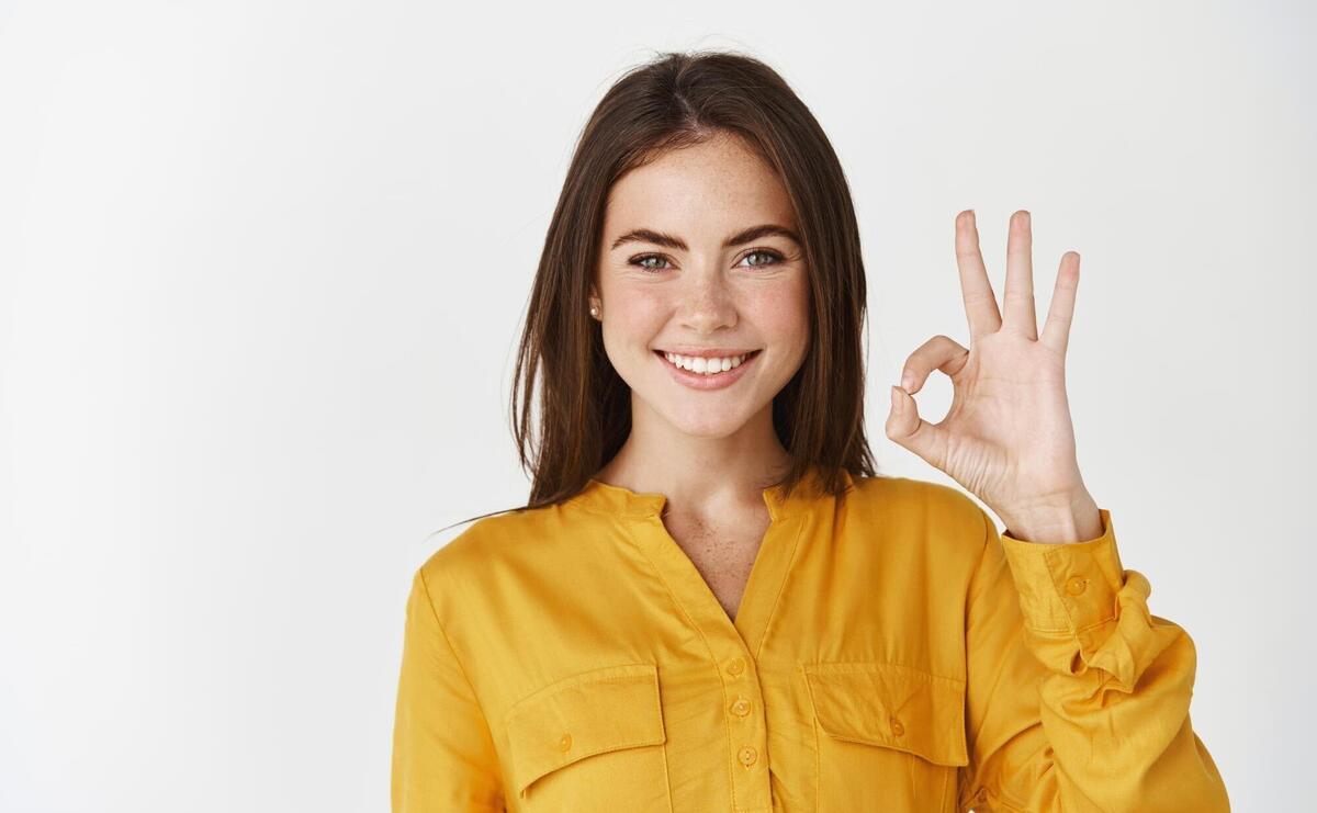 Young confident woman smiling, showing okay gesture, saying yes or alright and standing on white wall