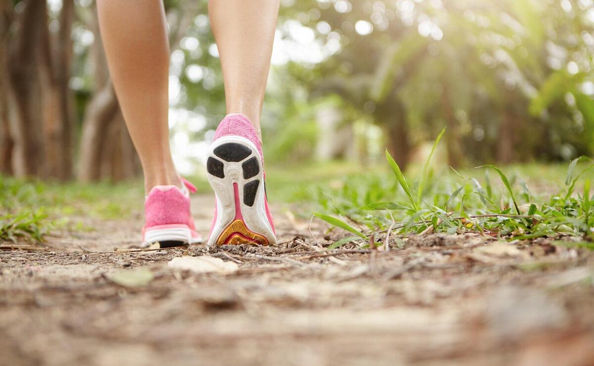 Cropped shot of athlete girl in pink running shoes hiking in forest on sunny day. Fit slim legs of sporty female in sneakers during jogging workout. Selective focus on sole.