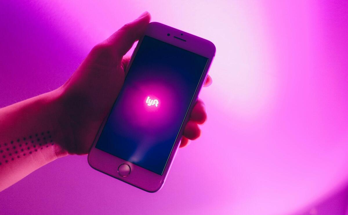 A hand holding a smartphone displaying the Lyft ridesharing app with a vivid pink background.