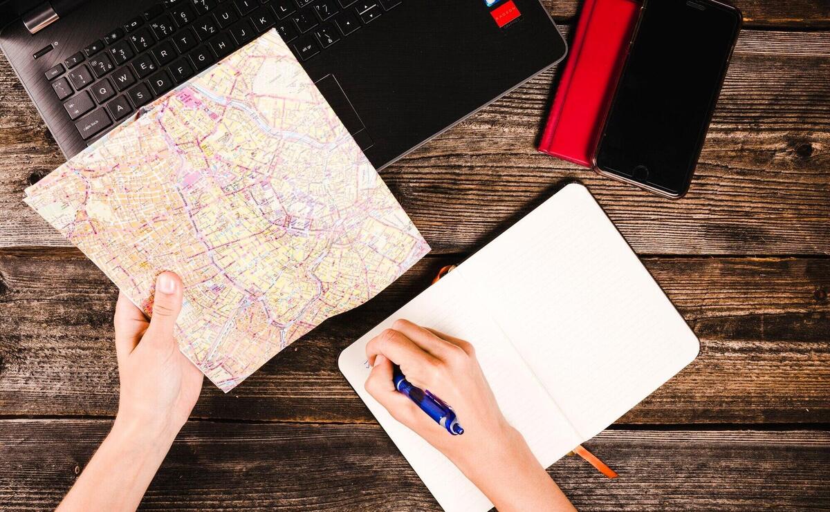 A person's hand writing in notepad while holding map over table