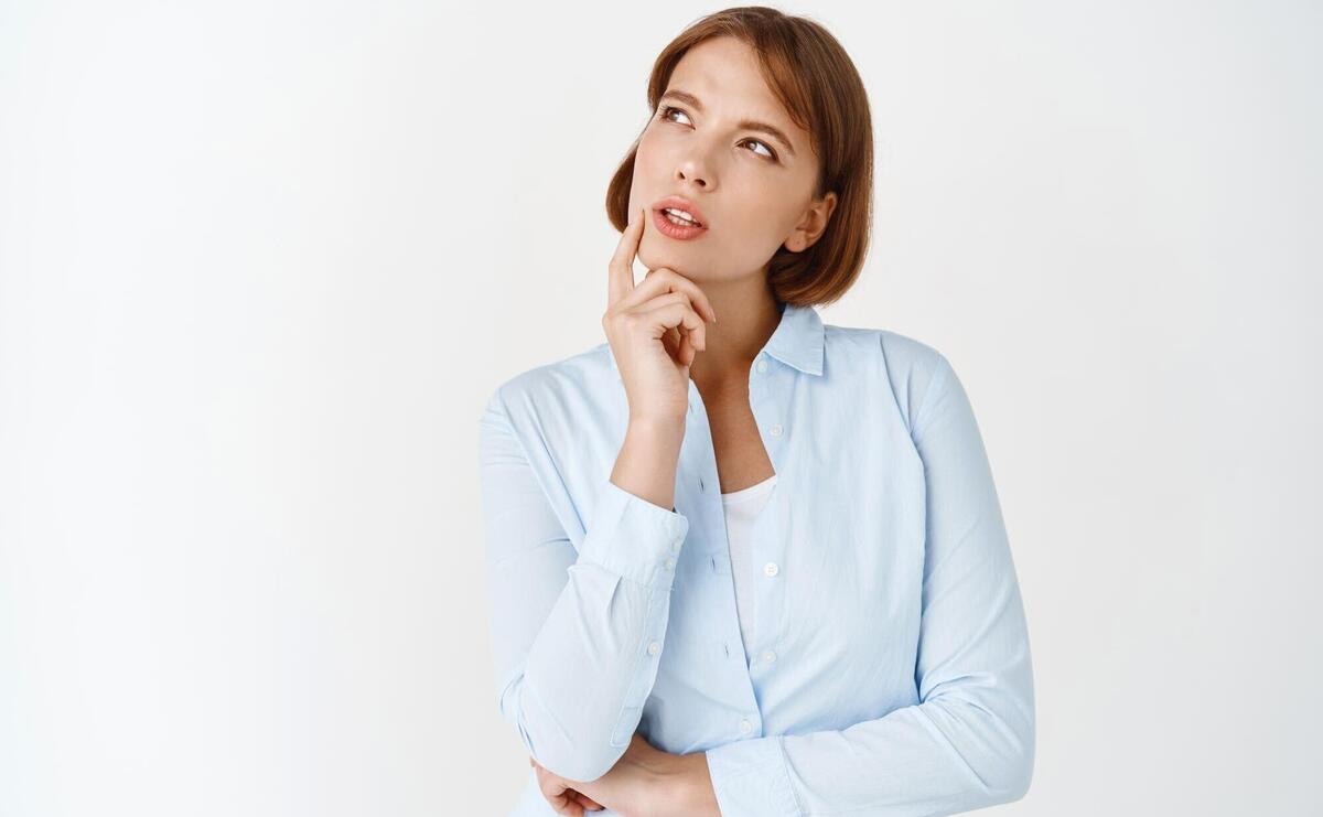 Portrait of young thoughtful woman looking aside at logo thinking and making choice pondering decision standing in blouse against white background