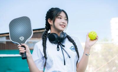 Smiling Asian woman holding a ball and paddle in outdoor setting. Urban sports vibe.