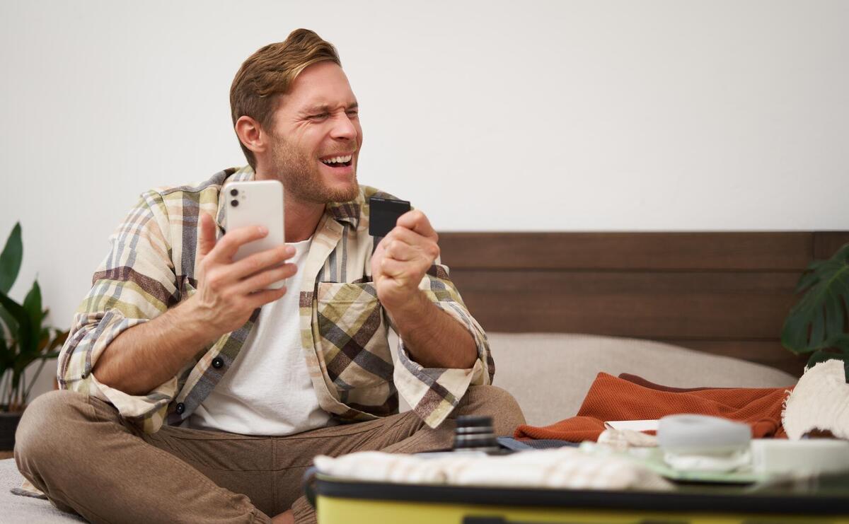 Cheerful male tourist man packing suitcase paying for tickets with credit card and smartphone app