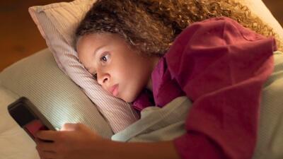 Side view of beautiful young girl lying in bed with her phone. Curly girl playing smart phone holding it in one hand moving finger on screen. Family, children leisure and using digital devices concept