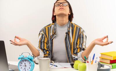 Peaceful young student girl wearing glasses sitting at desk meditating with closed eyes isolated on white background