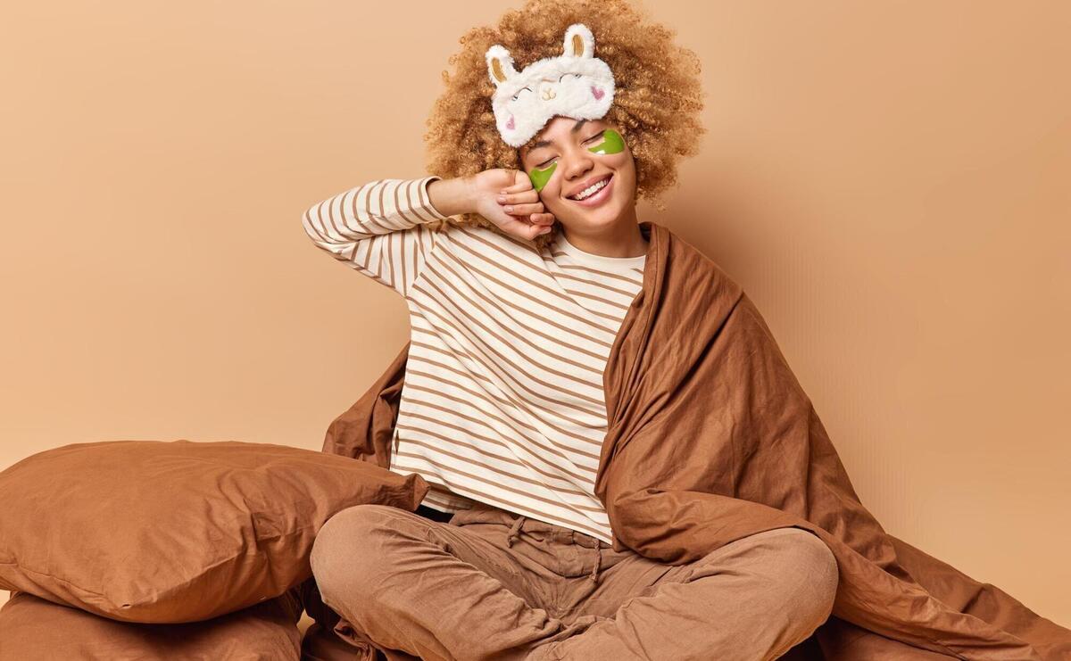 People and bed time concept Cheerful curly haired woman wears sleep mask and pajama sits crossed legs under blanket applies beauty hydrogel patches under eyes isolated over brown studio wall