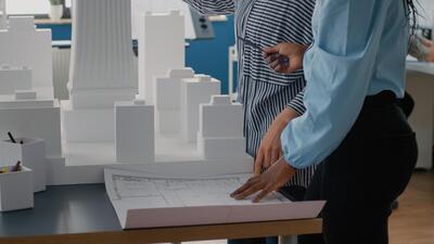 Close up of architects placing blueprints plan on table to work on urban project with building model. Team of women using layout print to design construction structure and development.